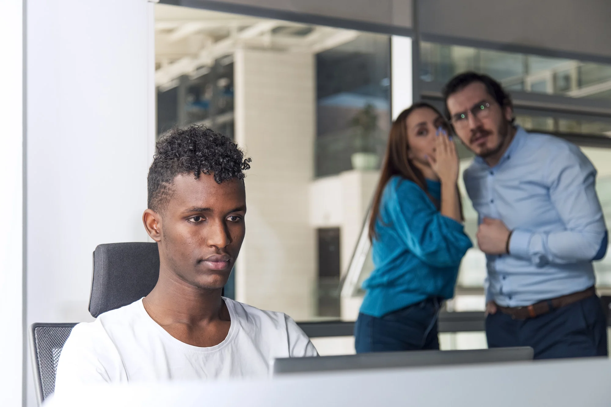 Black man is working on desk and there are two colleagues are gossiping at background behind of him